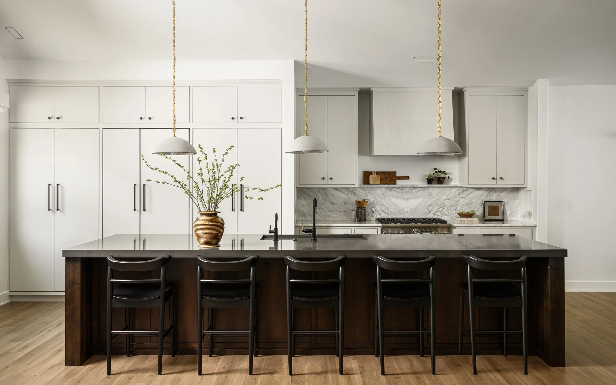 Modern kitchen with white cabinets, marble backsplash, and a large dark wood island with five black chairs. Decorative vase with branches on island, pendant lights, and minimal decor on backsplash.
