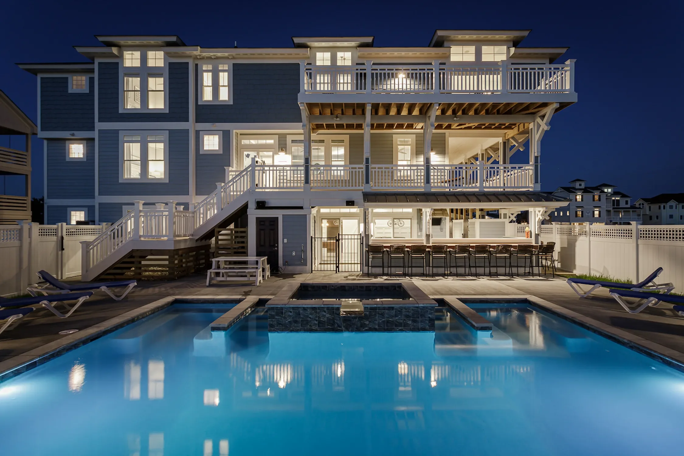 Nighttime view of a three-story house with illuminated windows and balconies, a backyard with a swimming pool, lounge chairs, and outdoor bar with stools.