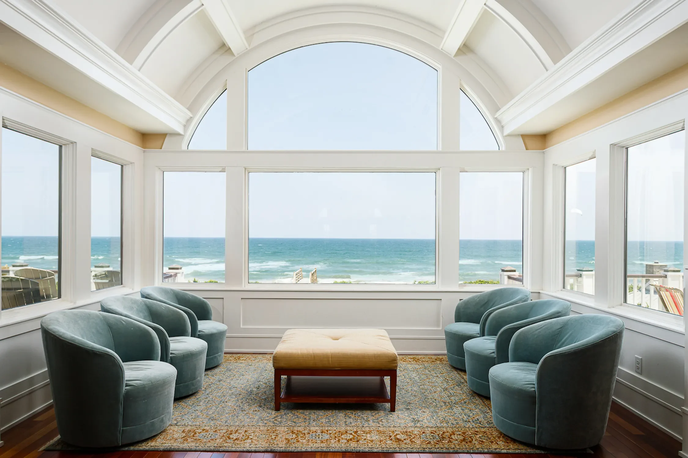 Seaside living room with large windows showing ocean view, six blue armchairs, a yellow ottoman, and a patterned rug.
