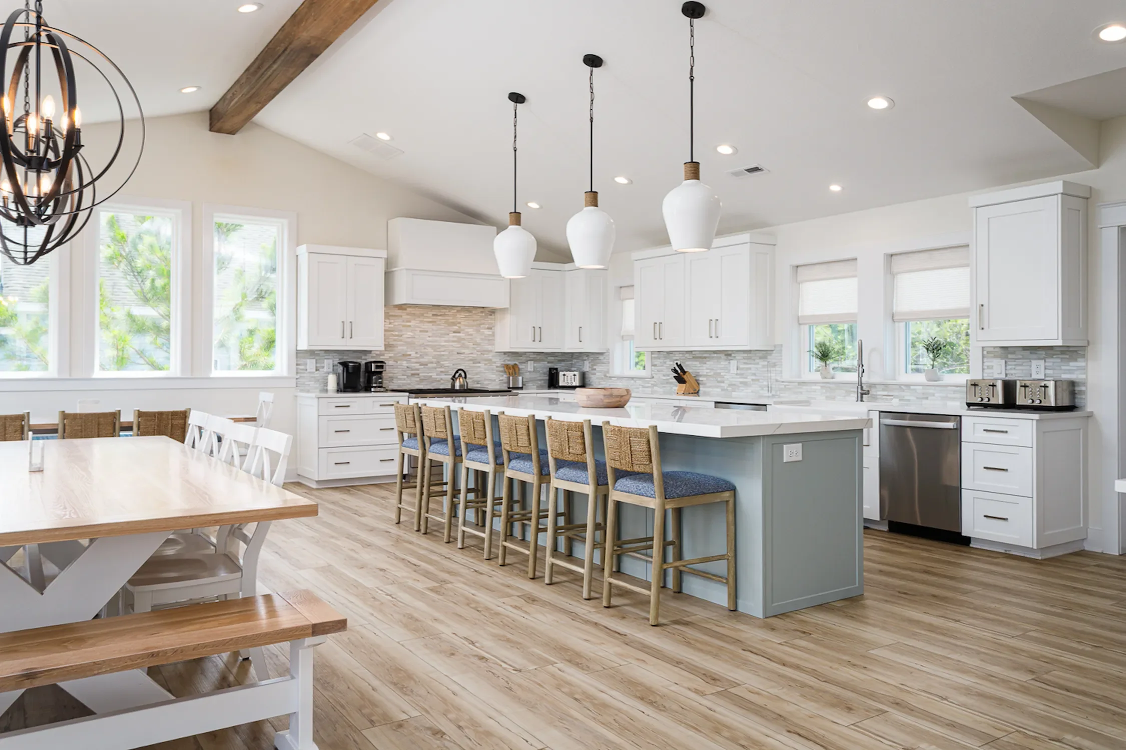 Open kitchen with white cabinets, a kitchen island with barstools, pendant lights, a dining table with chairs, large windows with blinds, and wooden flooring.