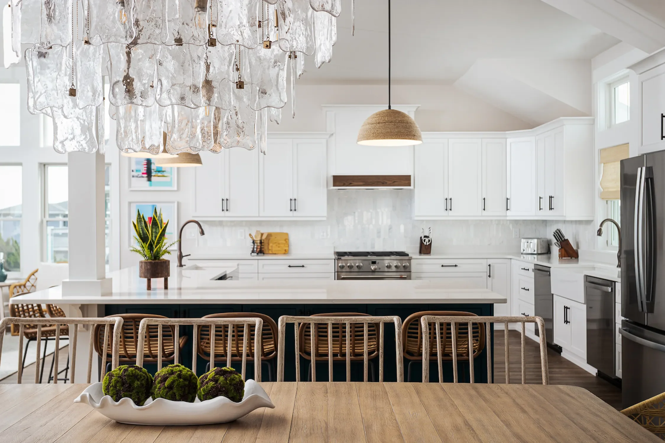 Modern kitchen with white cabinets, stainless steel appliances, a central island, wooden chairs, a wooden dining table with decorative green moss balls in a white dish, pendant light, and natural light from large windows.