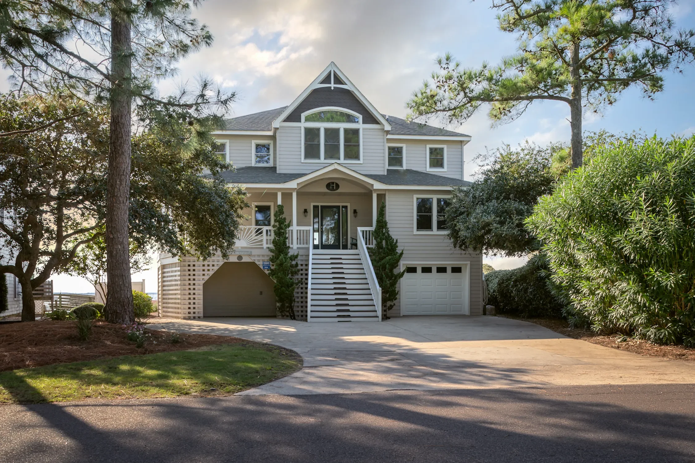 A large, multi-story house with a front porch, trees, and a driveway under a partly cloudy sky.