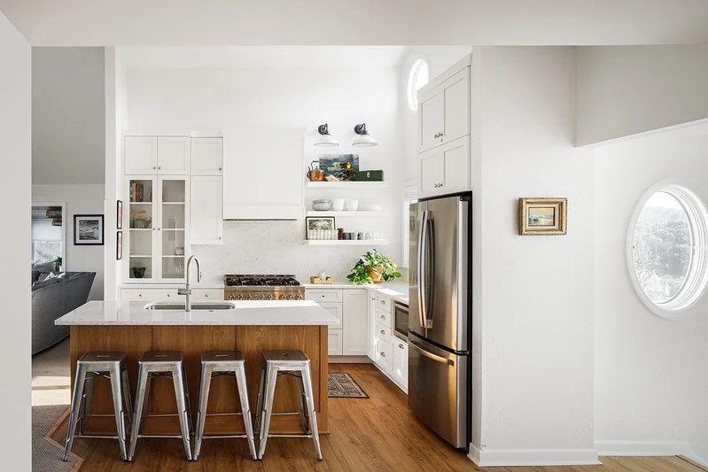 Modern kitchen with white cabinets, stainless steel refrigerator, and a wooden island with four metal stools. Open door and window provide natural light.