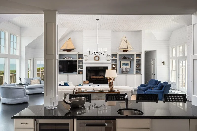 Bright living room with white walls and ceiling, large windows, and coastal decor, including model sailboats. There are blue and white armchairs, a white sofa, and a fireplace with built-in shelves. A kitchen island with a sink is in the foreground.