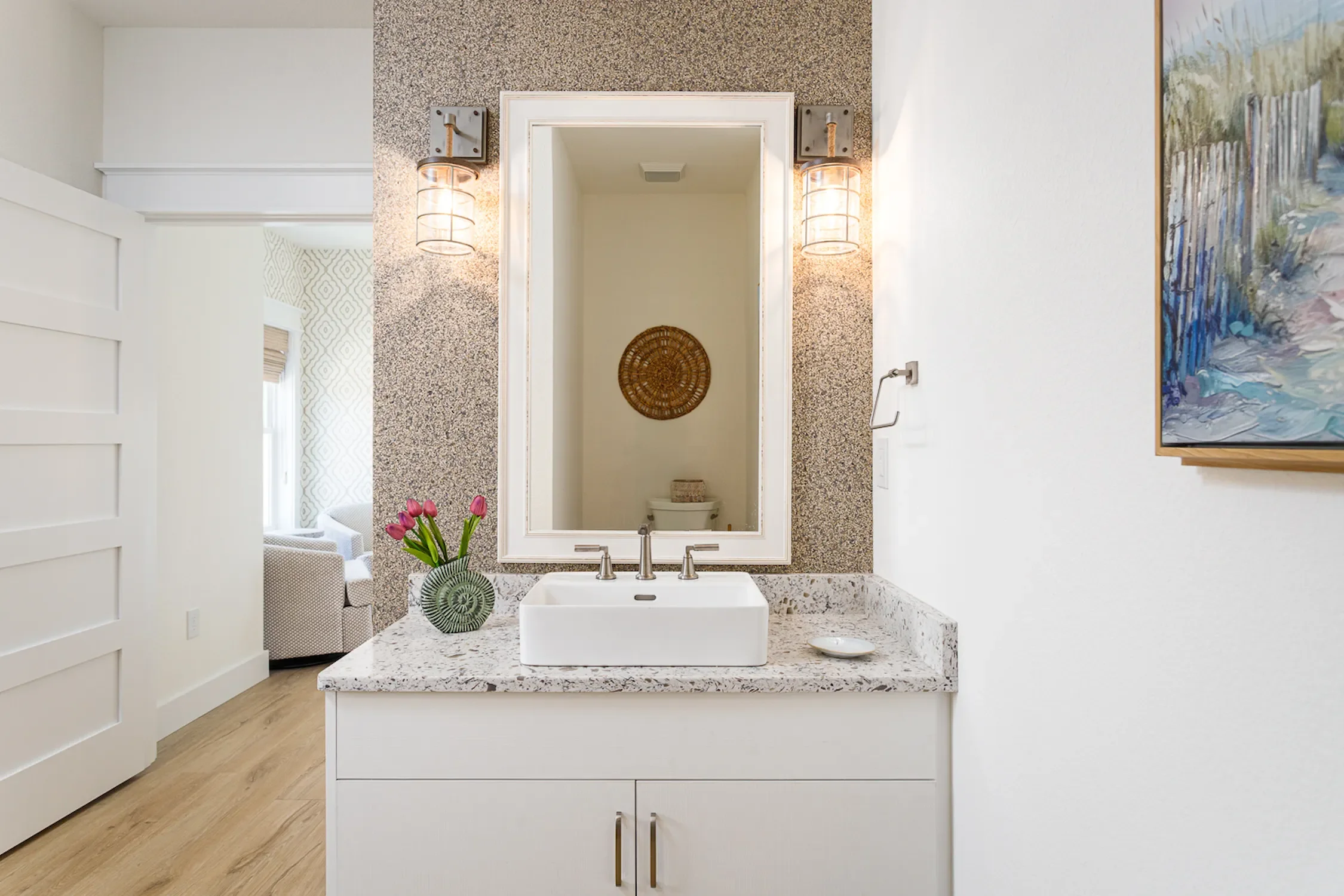 Bathroom vanity with a white cabinet, granite countertop, vase with pink tulips, and a rectangular mirror framed in white. Two wall-mounted light fixtures are on either side of the mirror. The scene reflects a toilet and woven wall decor in a neighbo