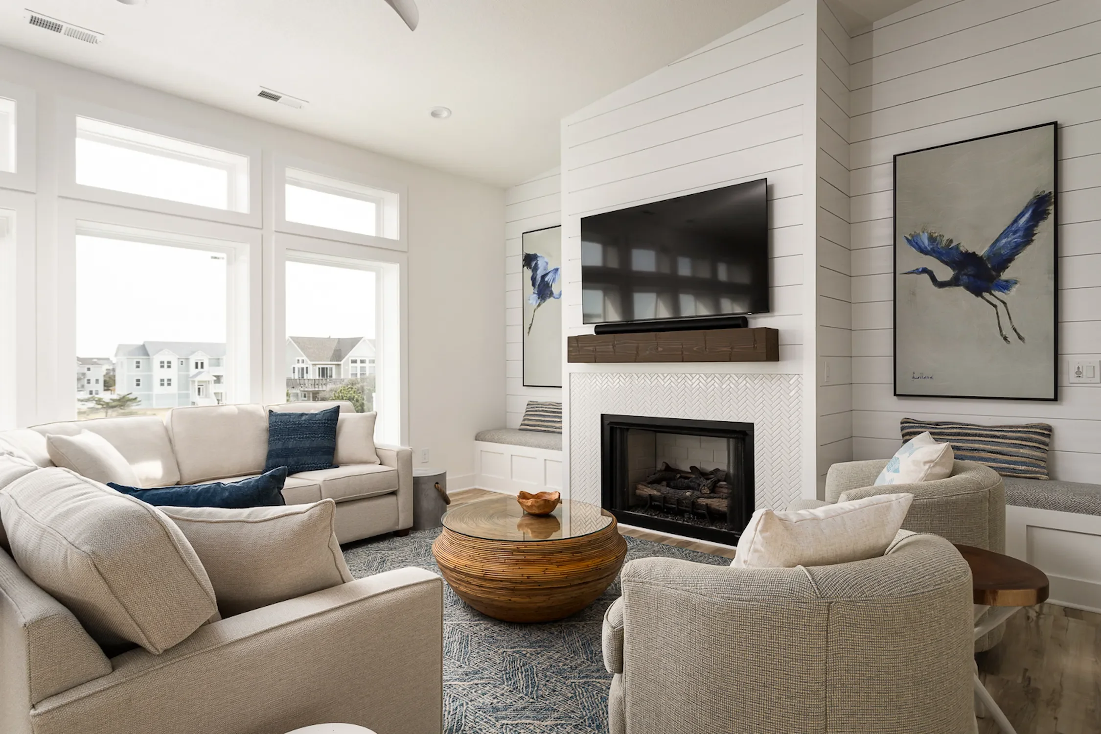 Modern living room with beige sofas, a round wooden coffee table, a fireplace, a wall-mounted TV, large windows, and wave art pictures with a blue heron painted on them.