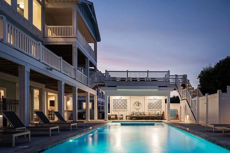 Outdoor swimming pool at sunset with lounge chairs, a two-story building with balconies, and a fenced patio area.