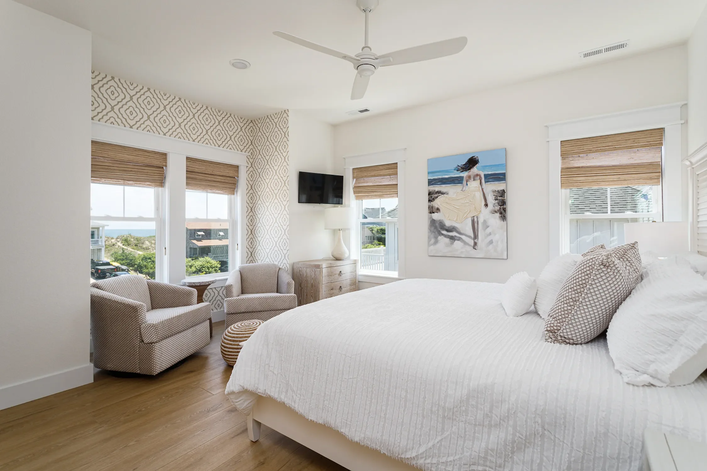 Bright bedroom with a large bed covered in white bedding, two patterned pillows, and three windows with blinds showing a coastal view. There are two patterned armchairs and a wood nightstand with a lamp, a mounted TV, and a piece of wall art depictin