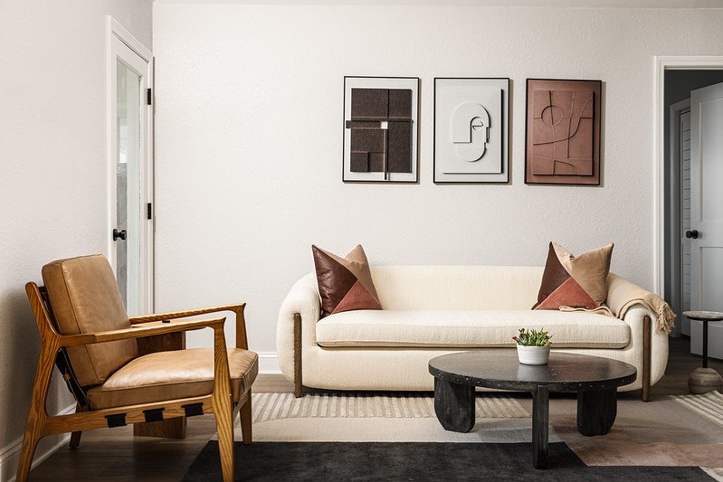 A living room featuring a beige sofa with brown and beige pillows, a round black coffee table with a small potted plant, a tan armchair with wooden arms, and three abstract art pieces on the white wall.