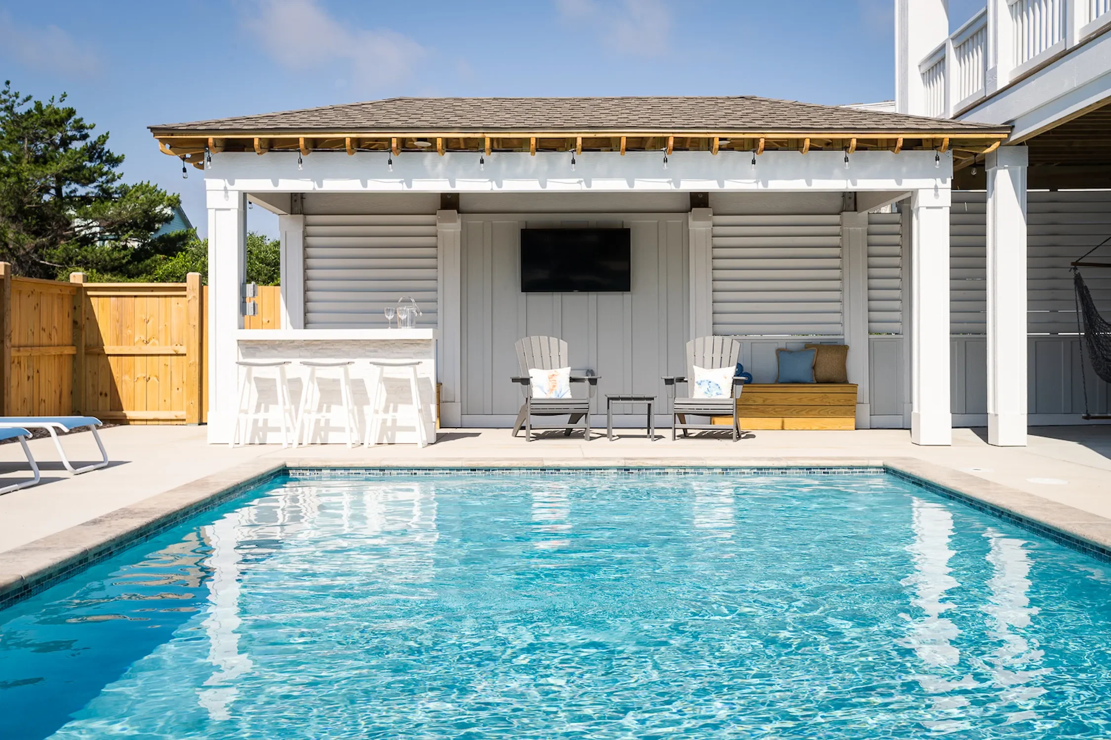 Swimming pool with clear blue water, surrounded by a concrete deck with lounge chairs, a poolside bar with white stools, and a covered patio area with two chairs, a bench with cushions, a small table, and a flat-screen TV mounted on the wall.