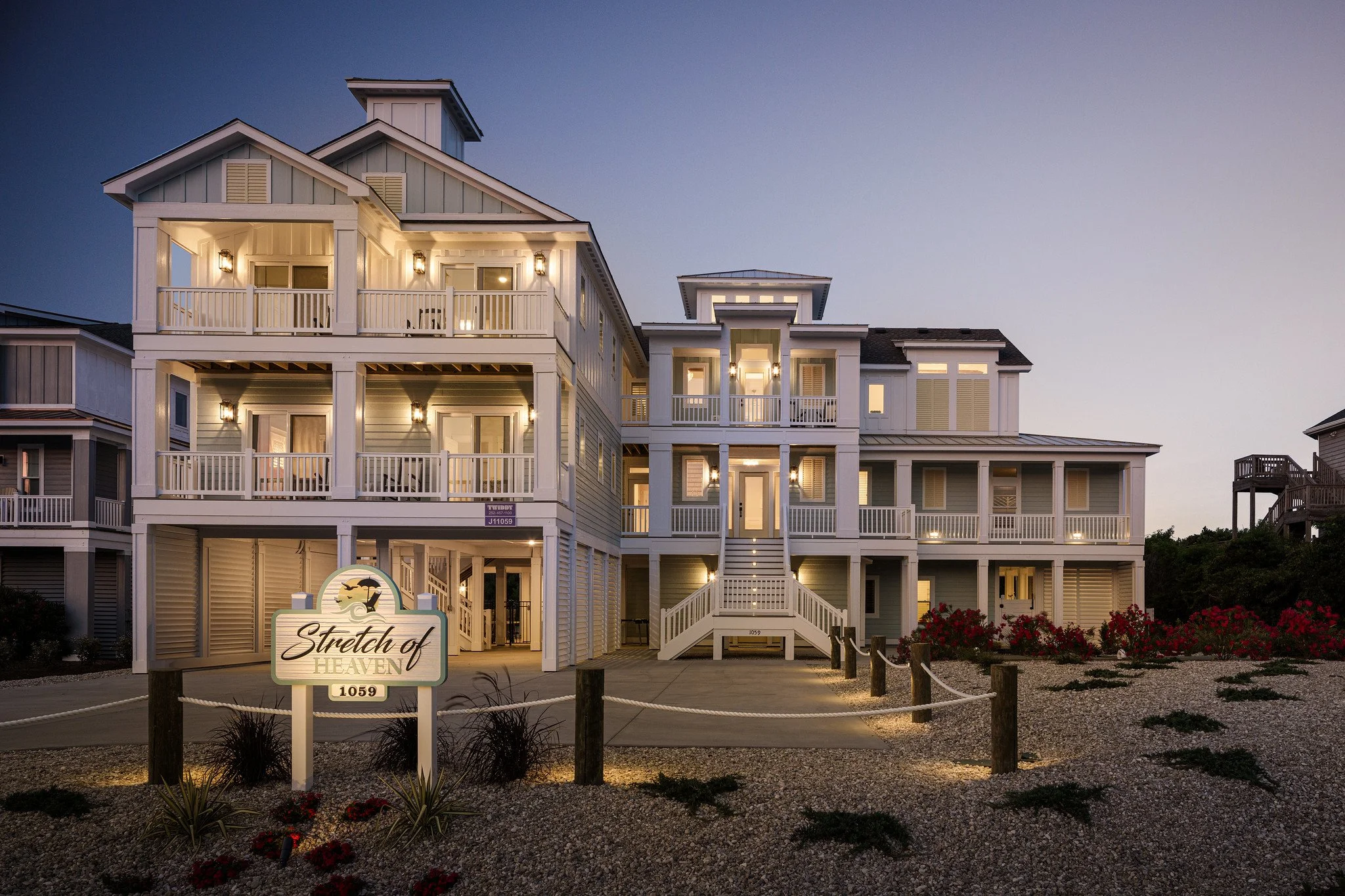 A large, multi-story beach house at dusk, with white exterior, balconies, and outdoor lighting, surrounded by a gravel courtyard and flower bushes, with a sign that reads 'Stretch of Heaven' and the number 1059.