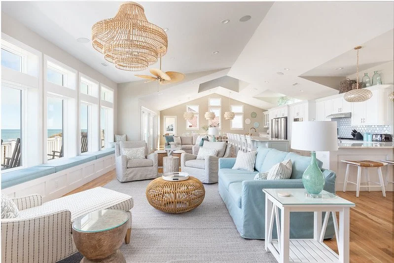 Bright living room with large windows, white walls, light blue and beige furniture, wicker chandelier, and an open kitchen in the background.
