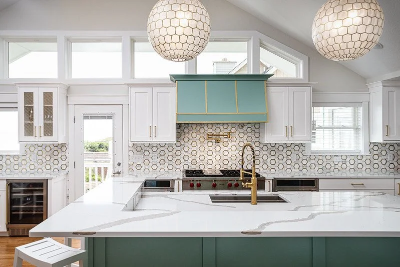 Modern kitchen with white cabinets, a marble island, and a hexagonal tile backsplash. Two large pendant lights hang from the ceiling, and a teal range hood is above the stove. Gold fixtures are also visible.