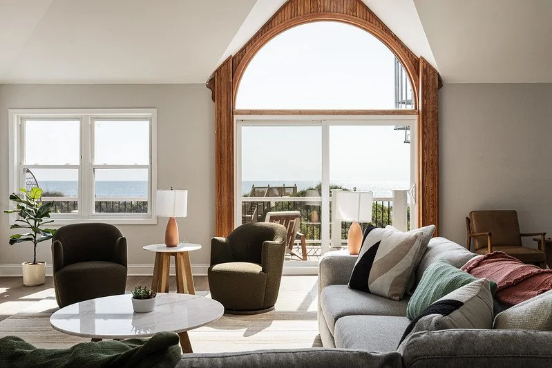 Living room with large arched window overlooking outdoor patio and ocean view, two green armchairs, a grey sofa with pillows, side table with lamp, and potted plant.