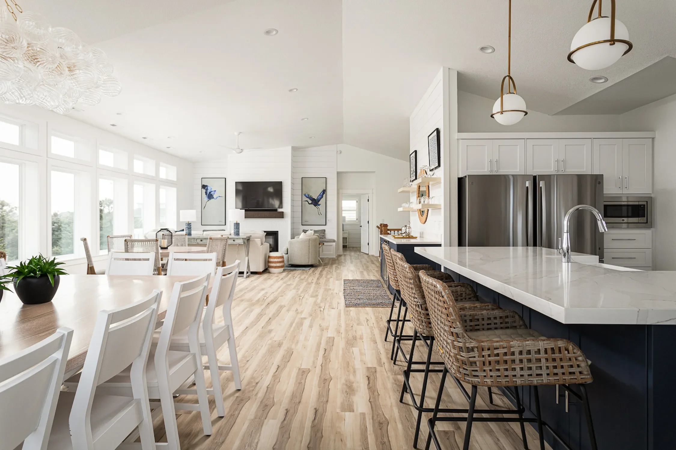 Open-concept living and kitchen area with white walls, large windows, wooden flooring, and modern furnishings. The kitchen has a white island with wicker chairs, stainless steel refrigerator, microwave, and white cabinetry. The living area features a