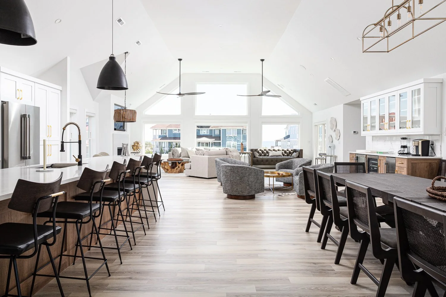 Open-concept living space with a kitchen island, bar stools, dining area, and a cozy seating area with sofas and chairs, large windows letting in natural light.