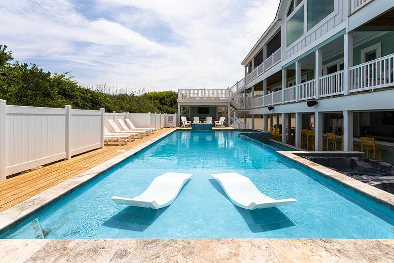 A swimming pool with two white lounge chairs inside, surrounded by a wooden deck, with a white fence and a multi-story coastal building in the background.
