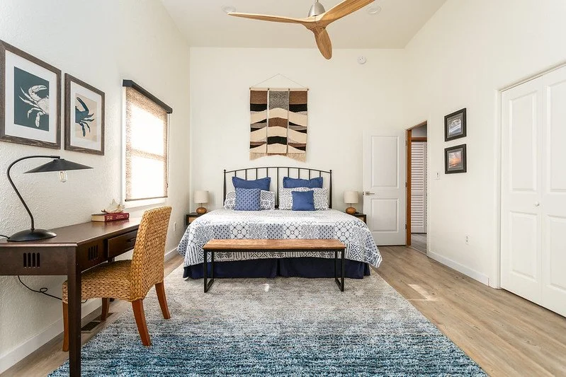 A bedroom with a bed, blue pillows, wall art, a wooden desk with a lamp, and a woven chair.