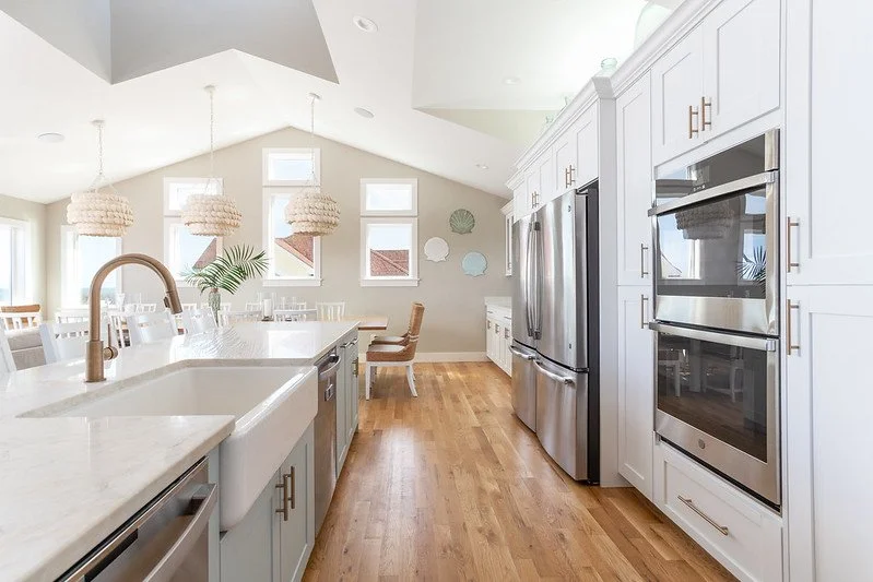 Bright, modern kitchen with white cabinets, stainless steel appliances, wooden flooring, and a large island with a sink. There are three pendant lights and multiple windows letting in natural light.