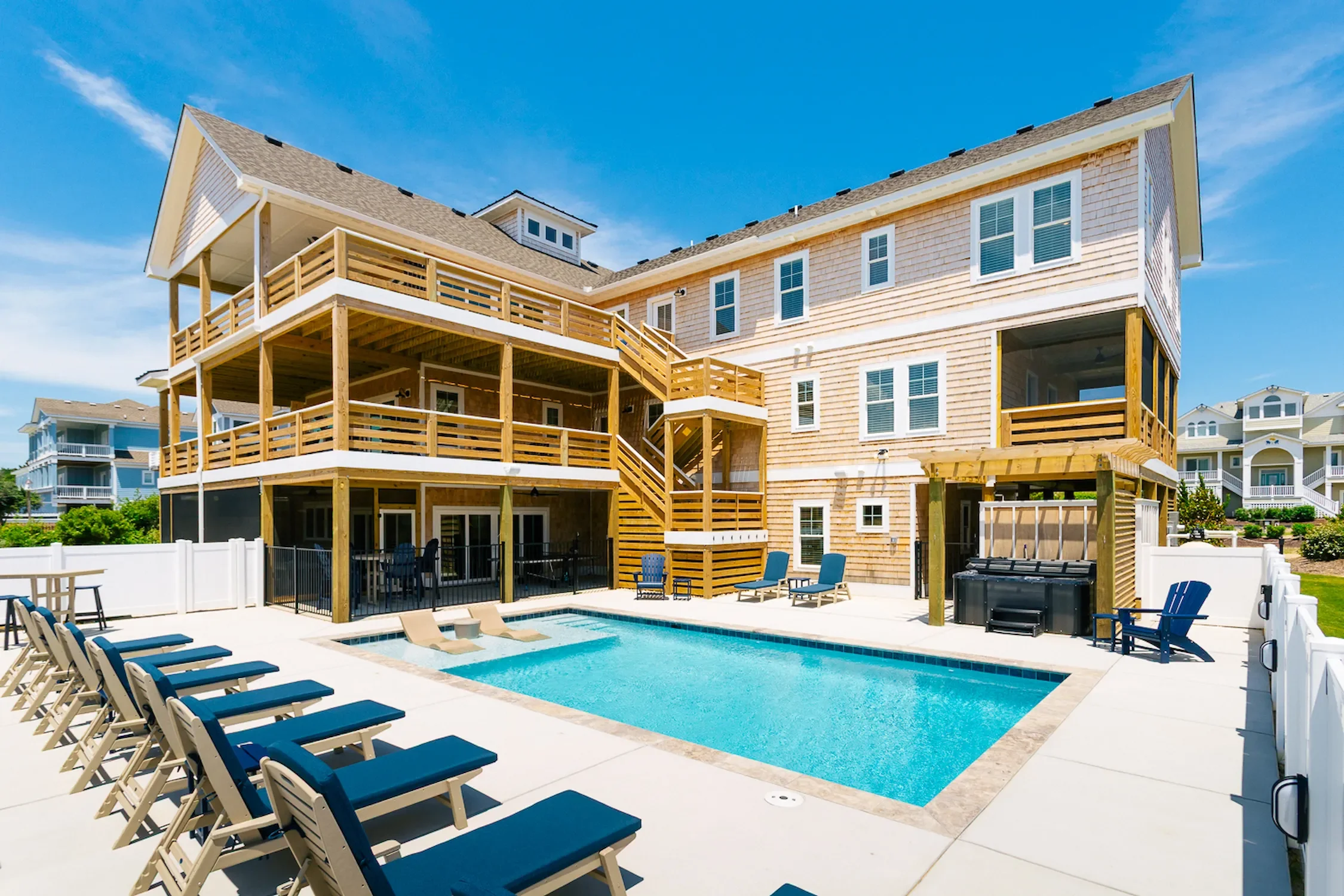 Large backyard with a swimming pool, several lounge chairs, and a multi-story house with a wooden deck and stairs, white fence, and neighboring houses in the background.