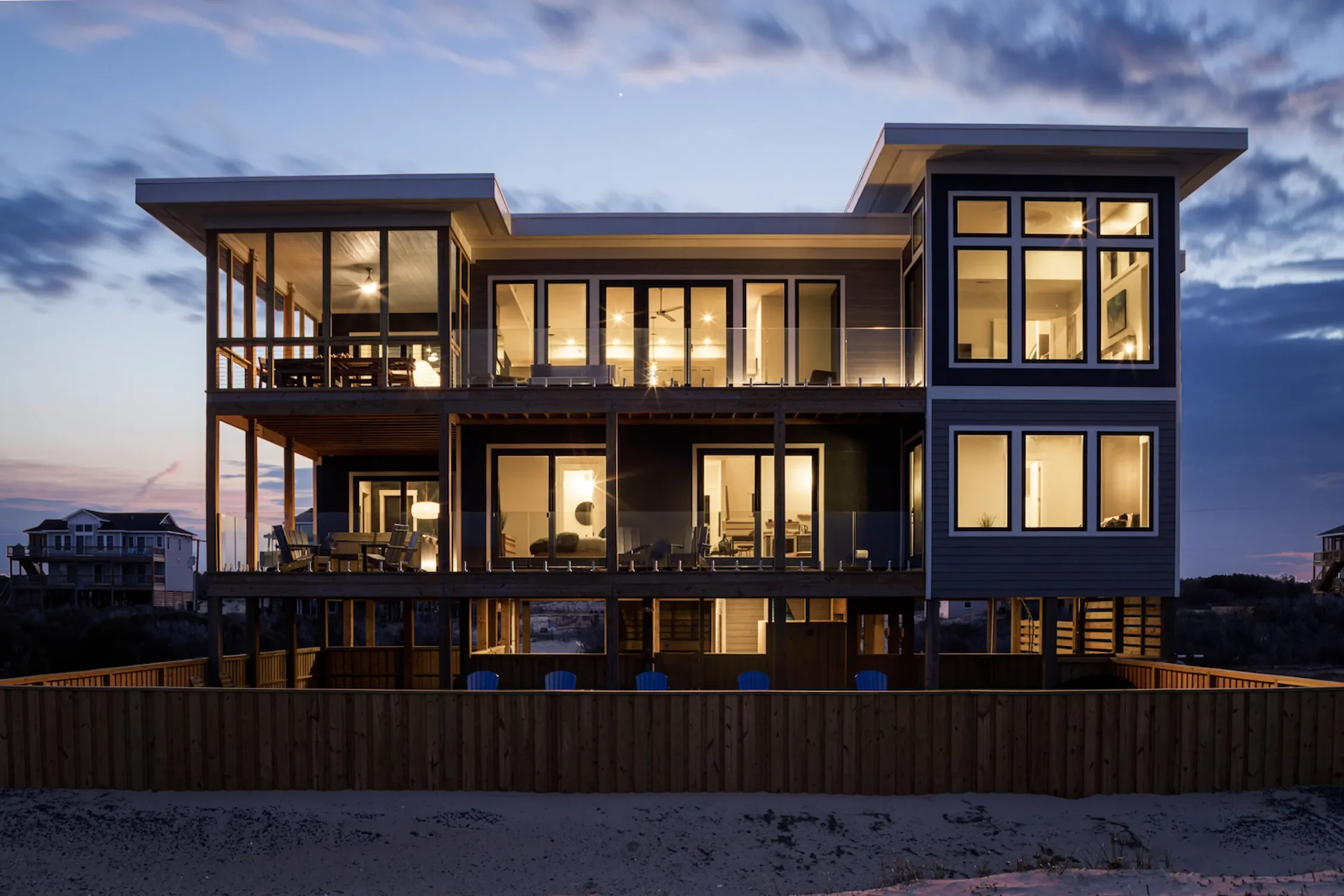Three-story modern house with large glass windows and balconies, illuminated from the inside, at dusk, with a beach and other houses in the background.