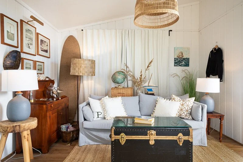 Living room with white paneled walls, a light gray sofa with white and patterned pillows, a black storage trunk coffee table, wooden side tables with lamps, and wall art including framed pictures, a painting, and a small hanging sculpture.