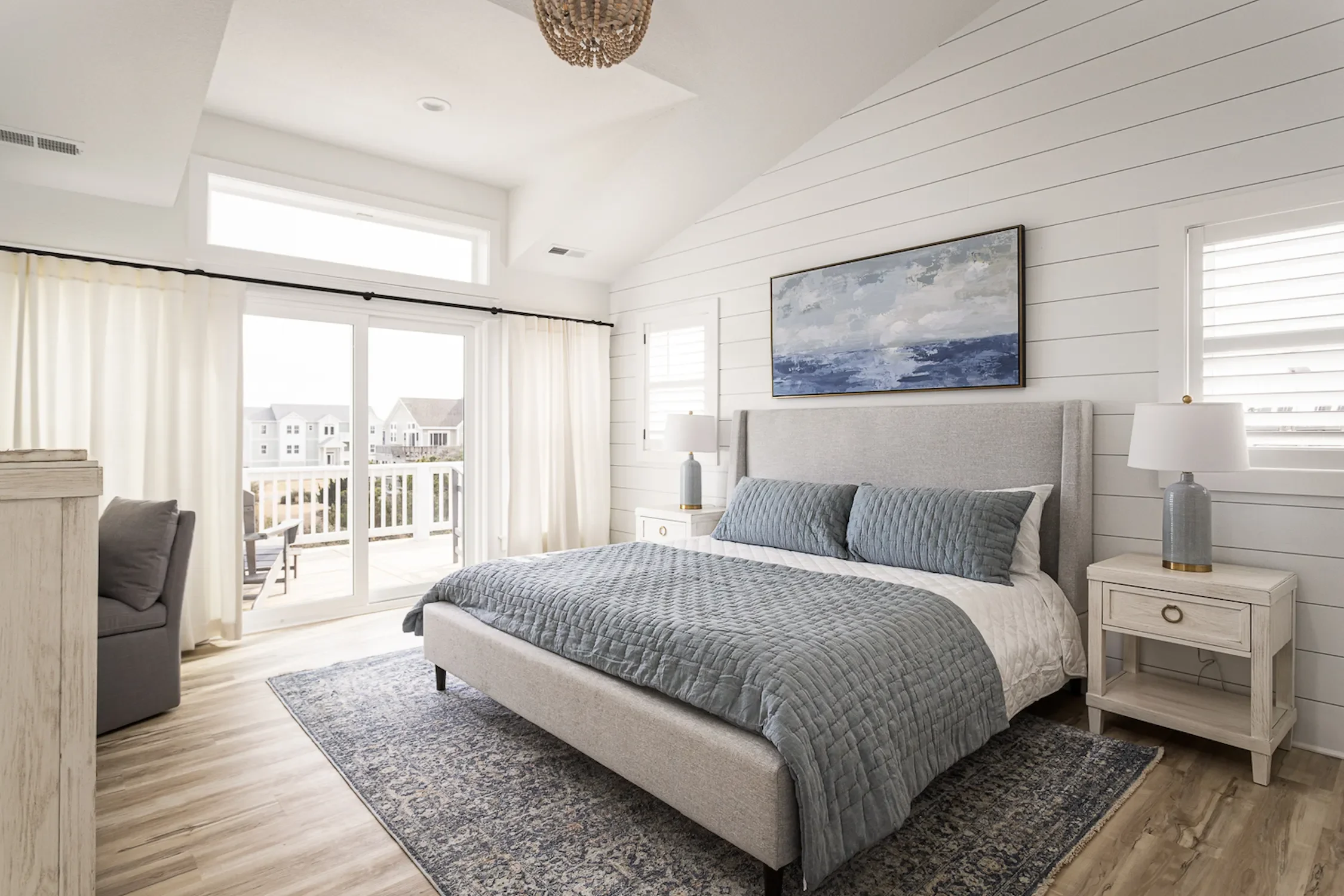 Bright bedroom with sliding glass door leading to balcony, white curtains, and coastal decor featuring a gray upholstered bed with gray bedding, matching pillows, two white nightstands with lamps, wood flooring, and a painting of an ocean scene.