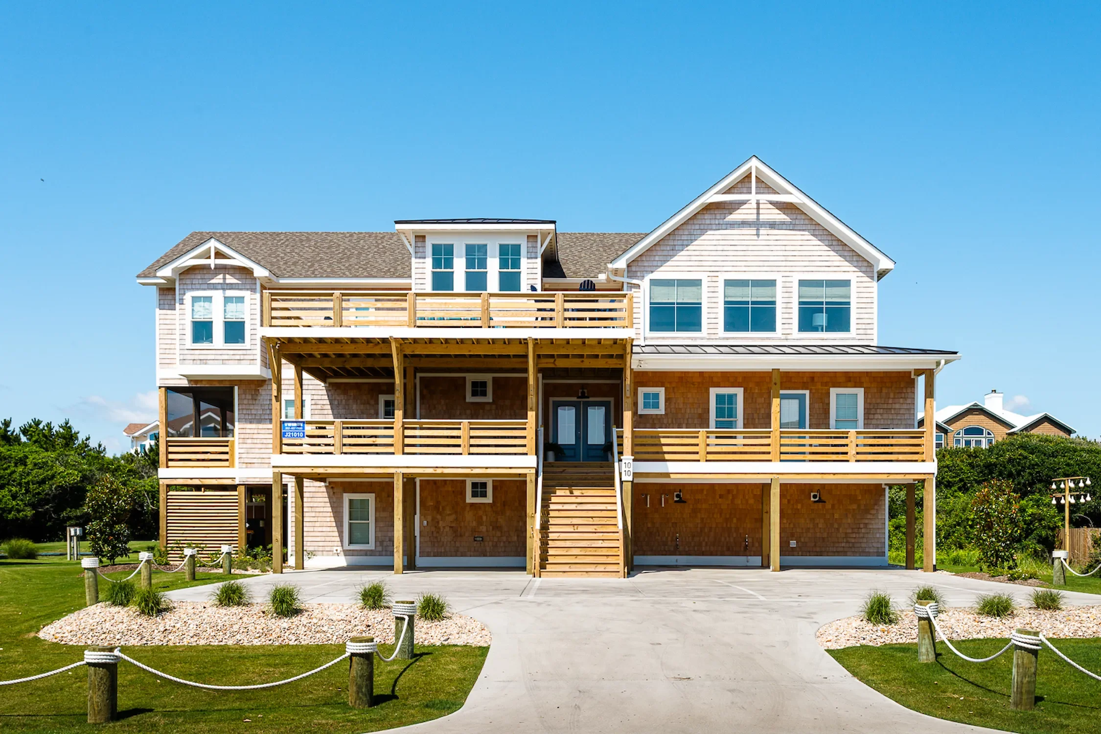 A large multi-story house with a wooden deck and stairs, beige siding, white trim, and several windows, set against a blue sky with some surrounding greenery.