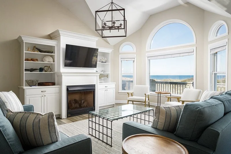 Living room with blue and striped sofas, a white fireplace with a mounted TV, white built-in shelves, a glass-topped coffee table, and a large window view of the ocean