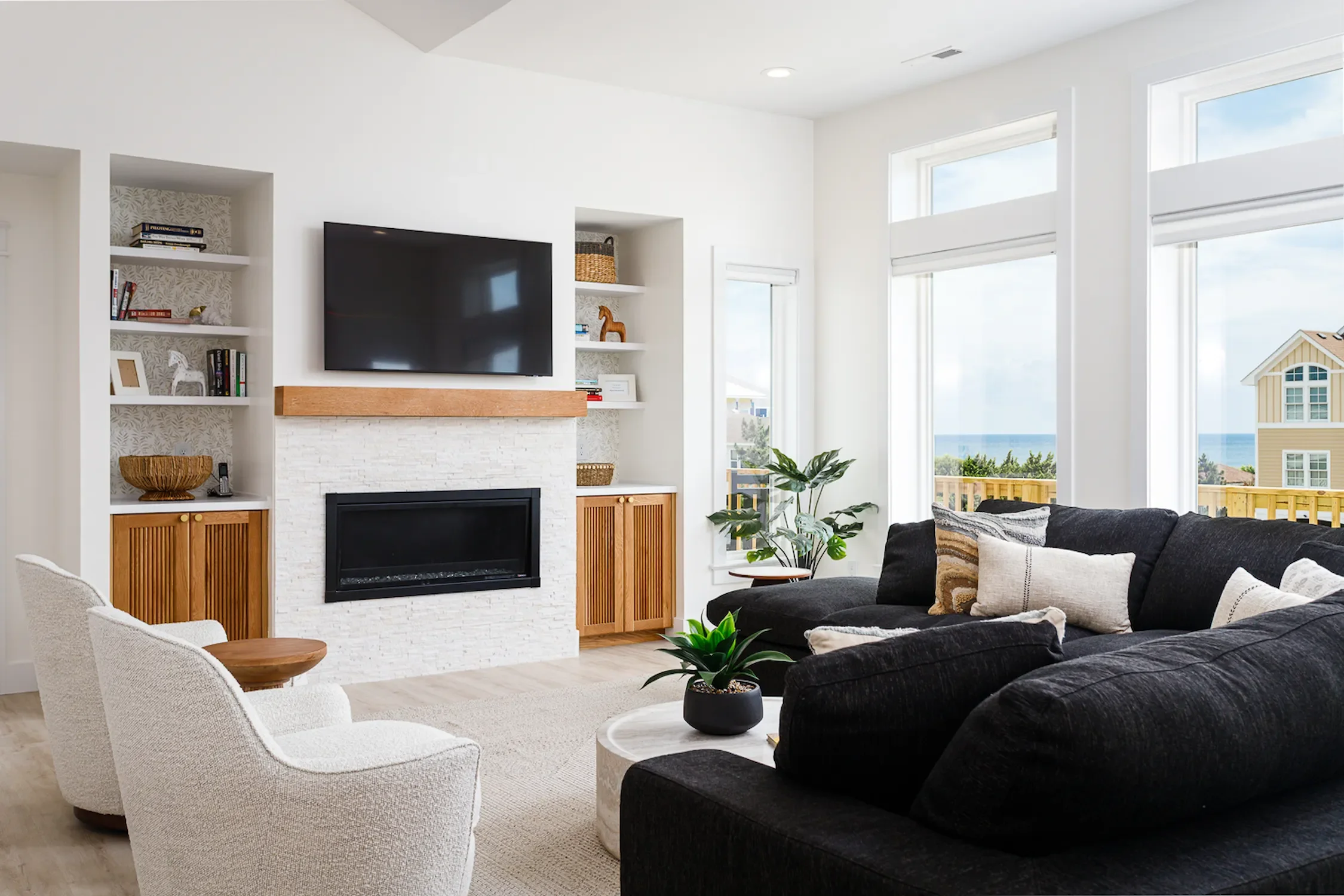 Living room with a white fireplace, a large flat-screen TV, built-in shelving, a black sectional sofa with beige and patterned pillows, white armchairs, a small round table with a plant, and large windows with a view of a beach and neighboring house.
