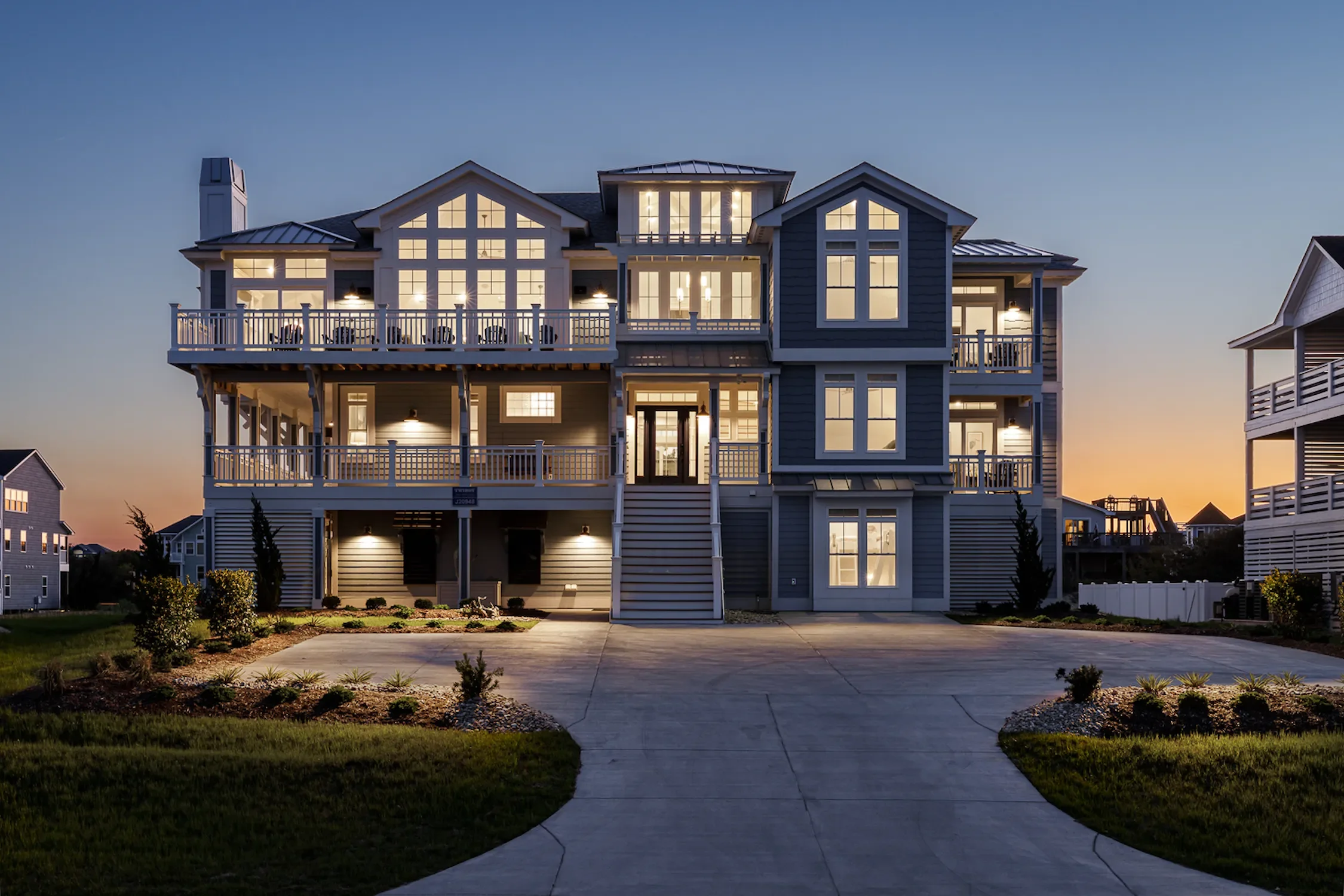 A large, multi-story house with blue siding and white trim, with multiple lit windows and balconies, illuminated at sunset or dusk.