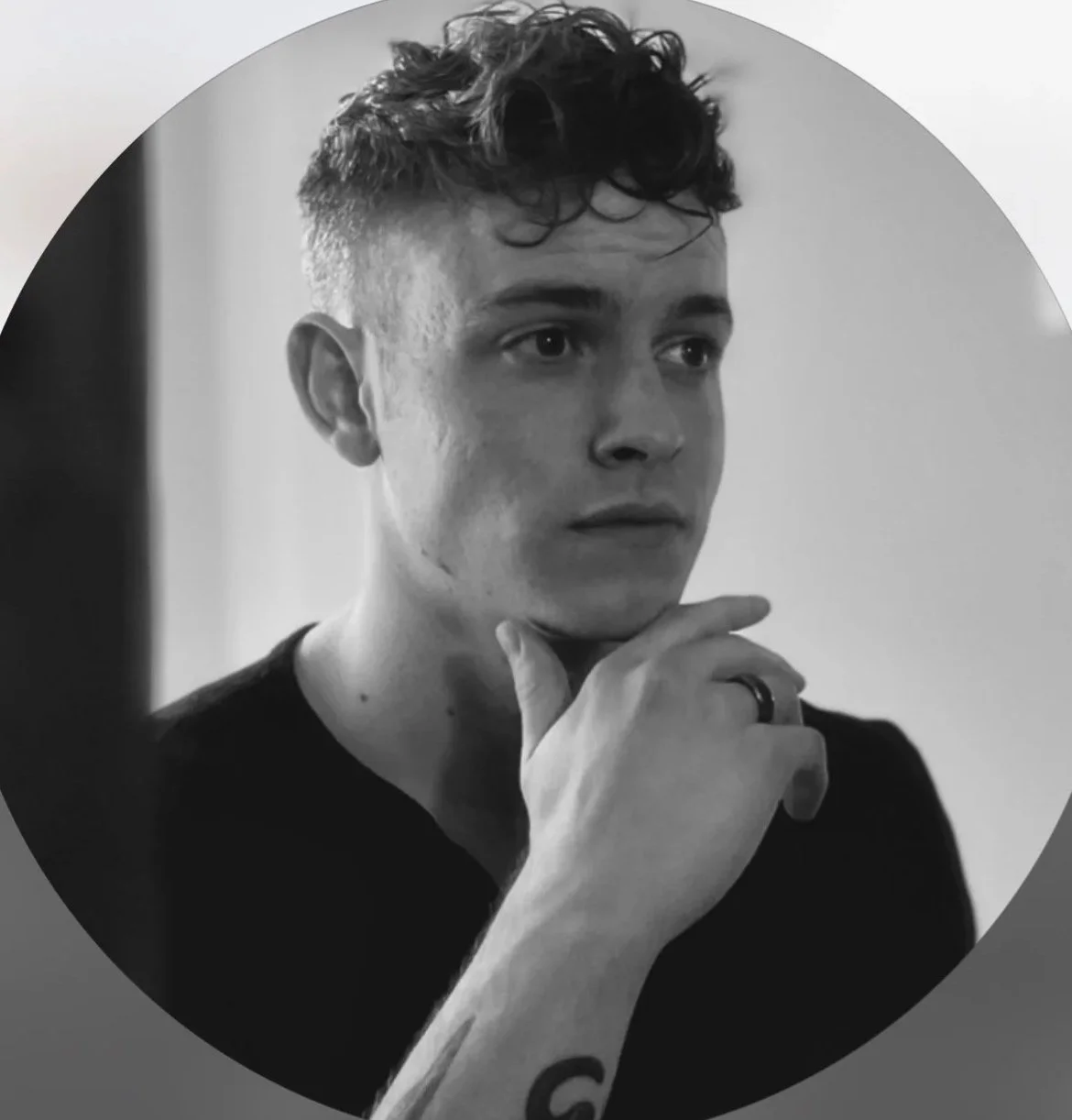 Black and white photo of a young man with short, curly hair, wearing a dark shirt, with a contemplative expression, resting his chin on his hand and looking off to the side.
