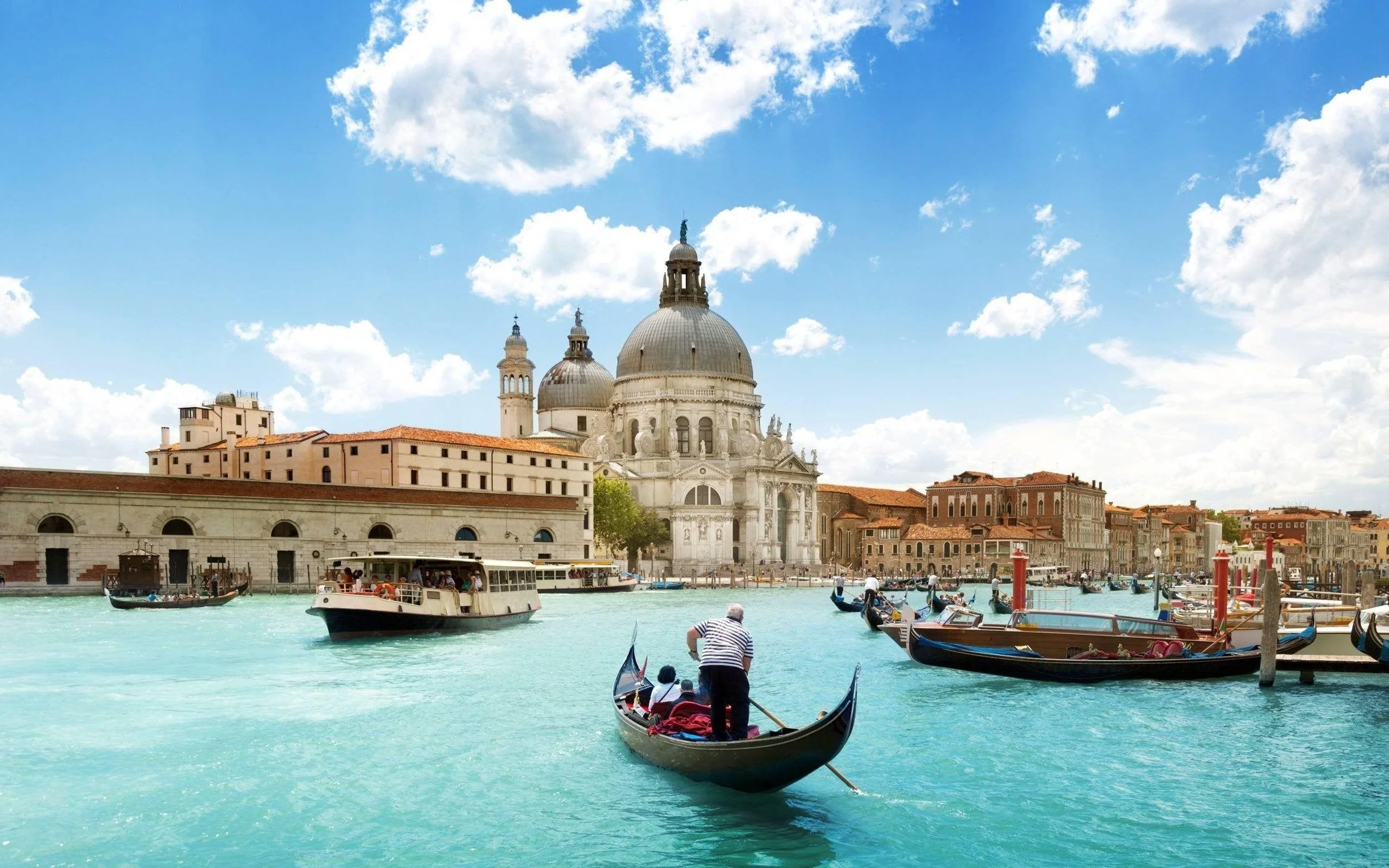 Gondola rides on turquoise waters with historical buildings and the Basilica di Santa Maria della Salute in Venice, Italy, under a partly cloudy sky.