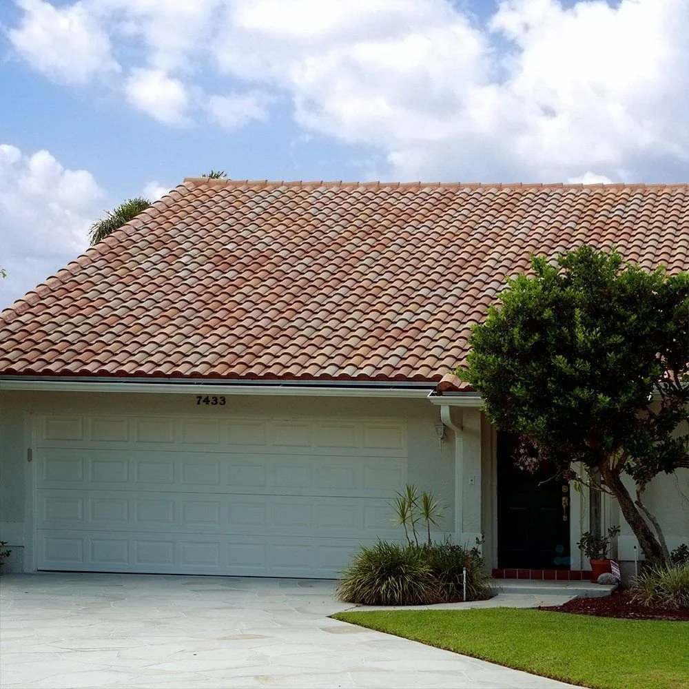 Front view of a house with a tiled roof, white garage door, and a tree near the entrance, under a partly cloudy sky.