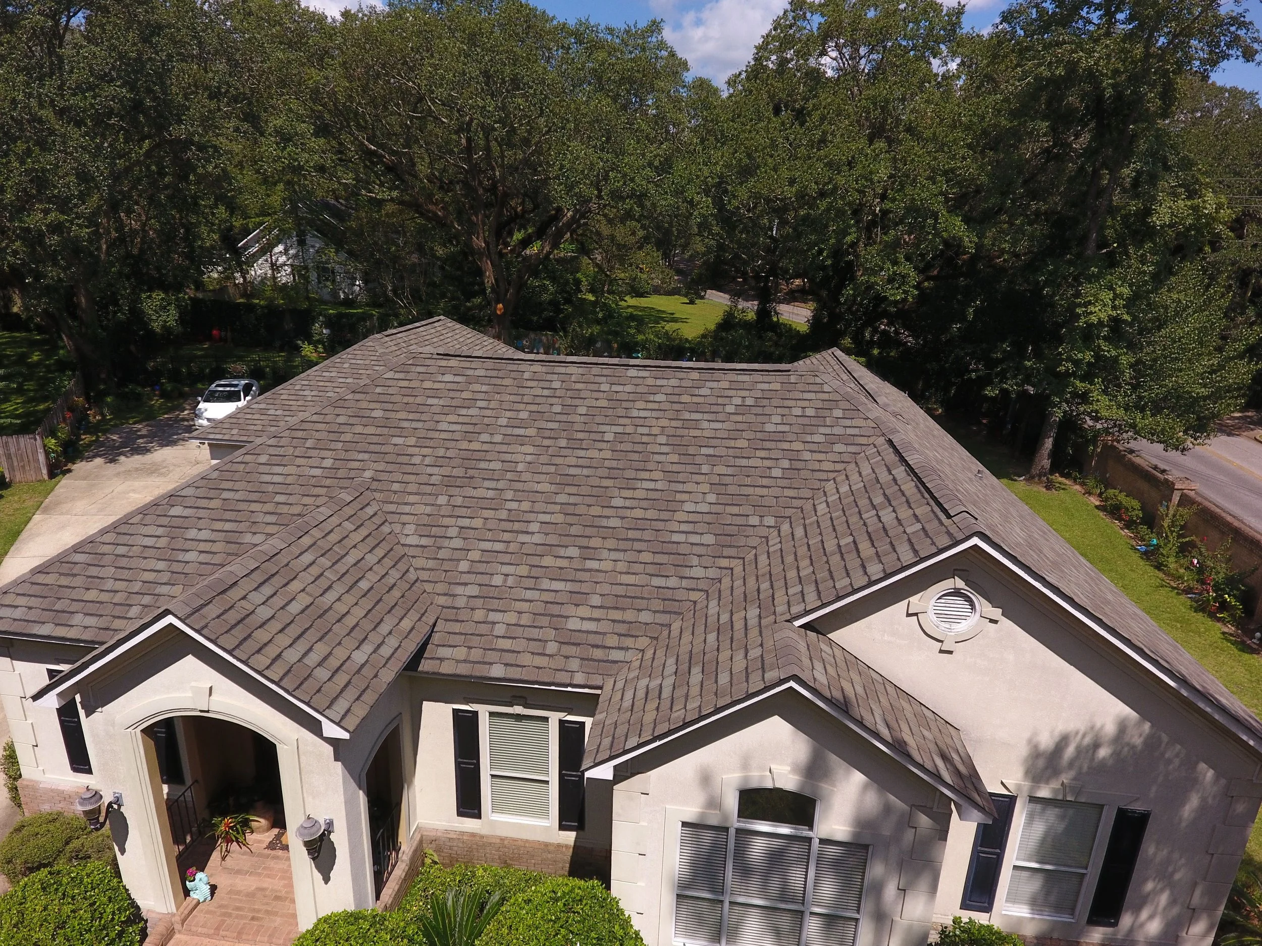 Aerial view of a house with a gray shingle roof, front porch, windows with black shutters, and a well-maintained lawn, surrounded by trees and a fence.