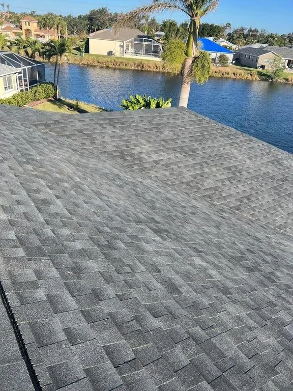 View of a grey shingled roof overlooking a lake with houses and palm trees in the background.