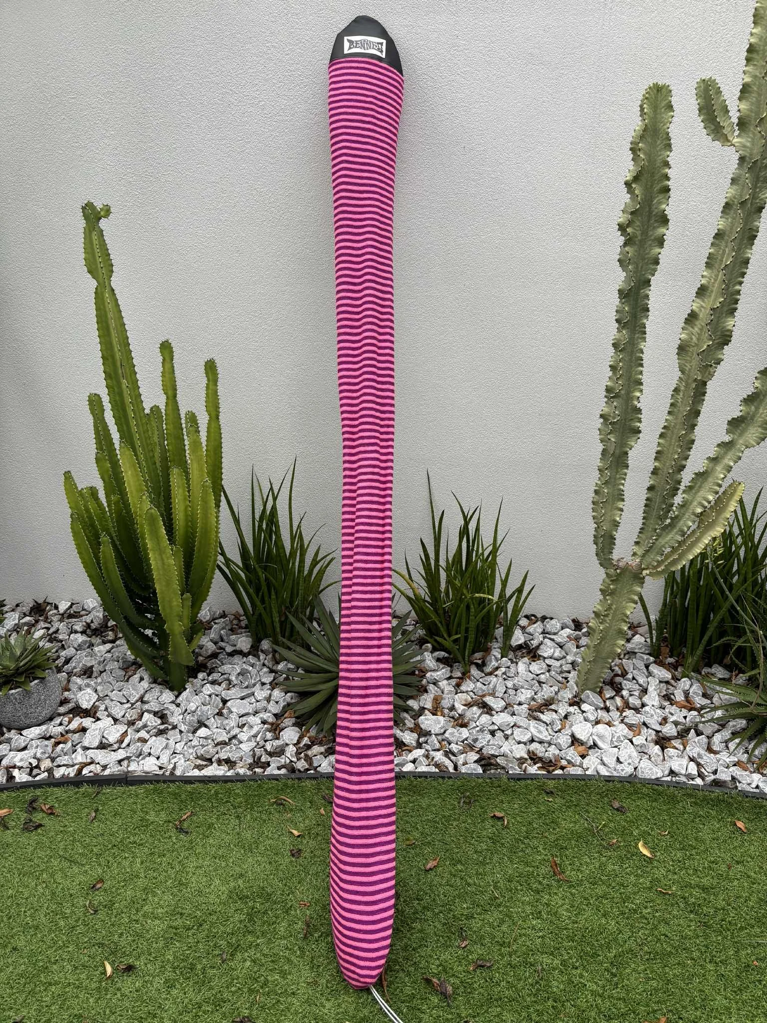 A pink and black striped fleece cover for fixed length surf ski paddle leaning against a wall, surrounded by desert plants and small rocks on the ground.