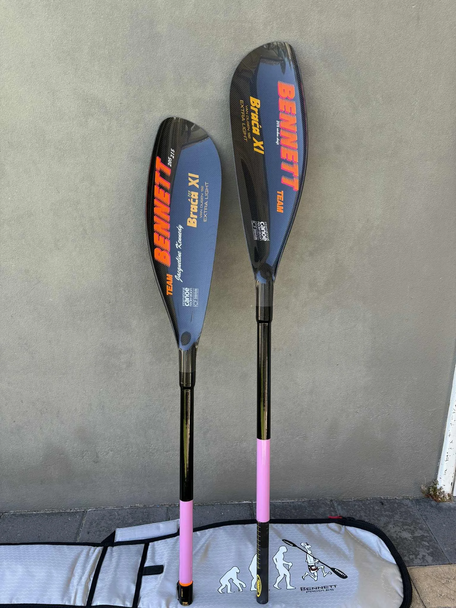 A Bennett Braca XV paddles with black shafts, pink grips, and blue blades with orange and yellow text, leaning against a gray wall above a Bennett paddle board.