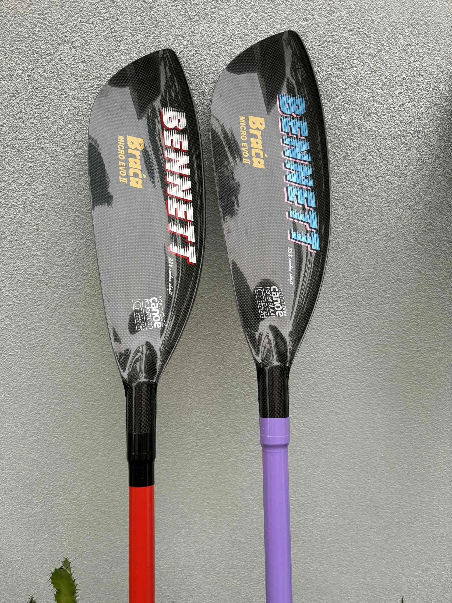 Two carbon fiber paddles with digital branding, one with a red handle and the other with a purple handle, hanging on a textured gray wall.