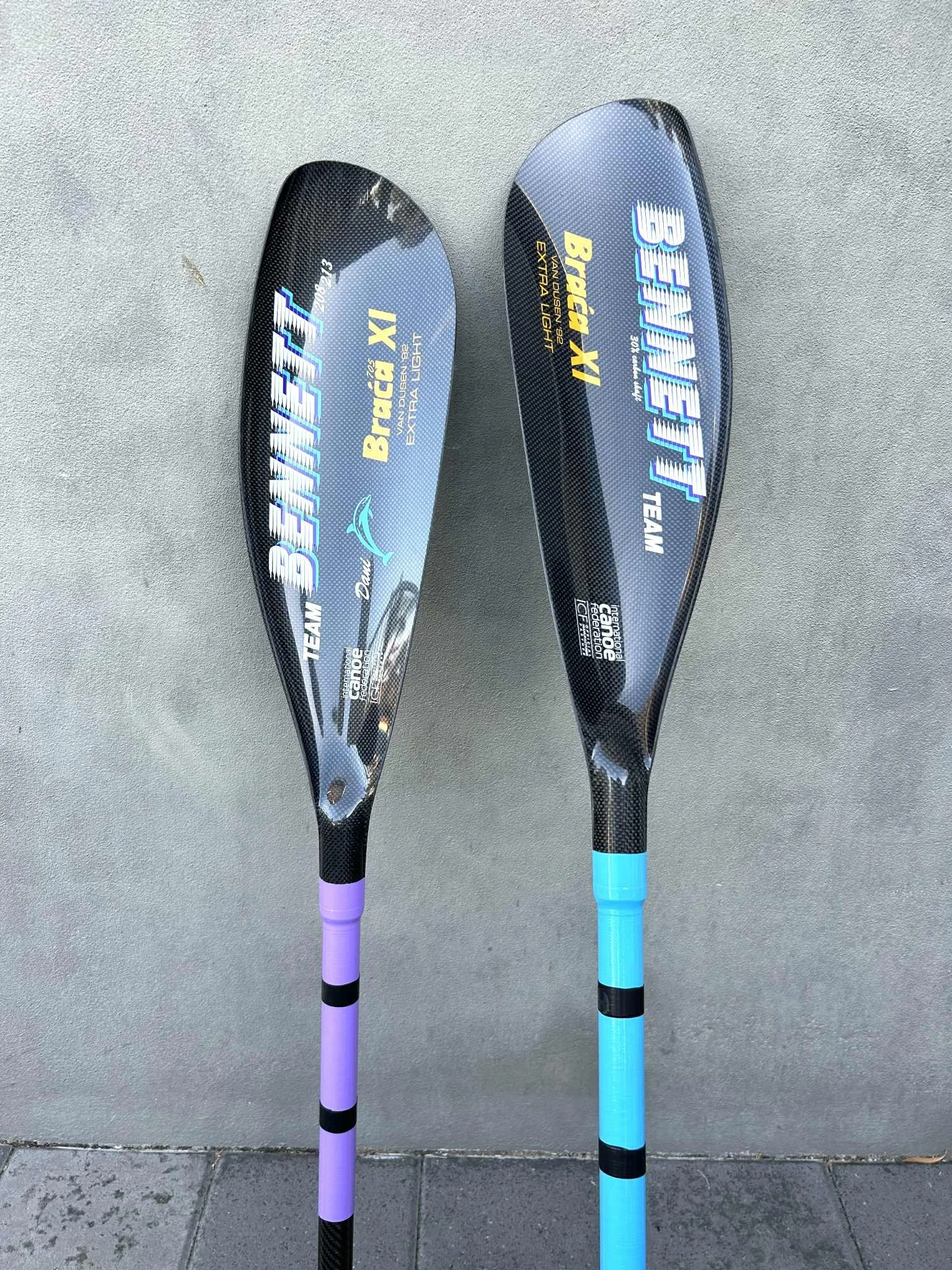 Two carbon fiber paddles with Team Bennett XI and extra light written on them, placed on a concrete ground against a wall.