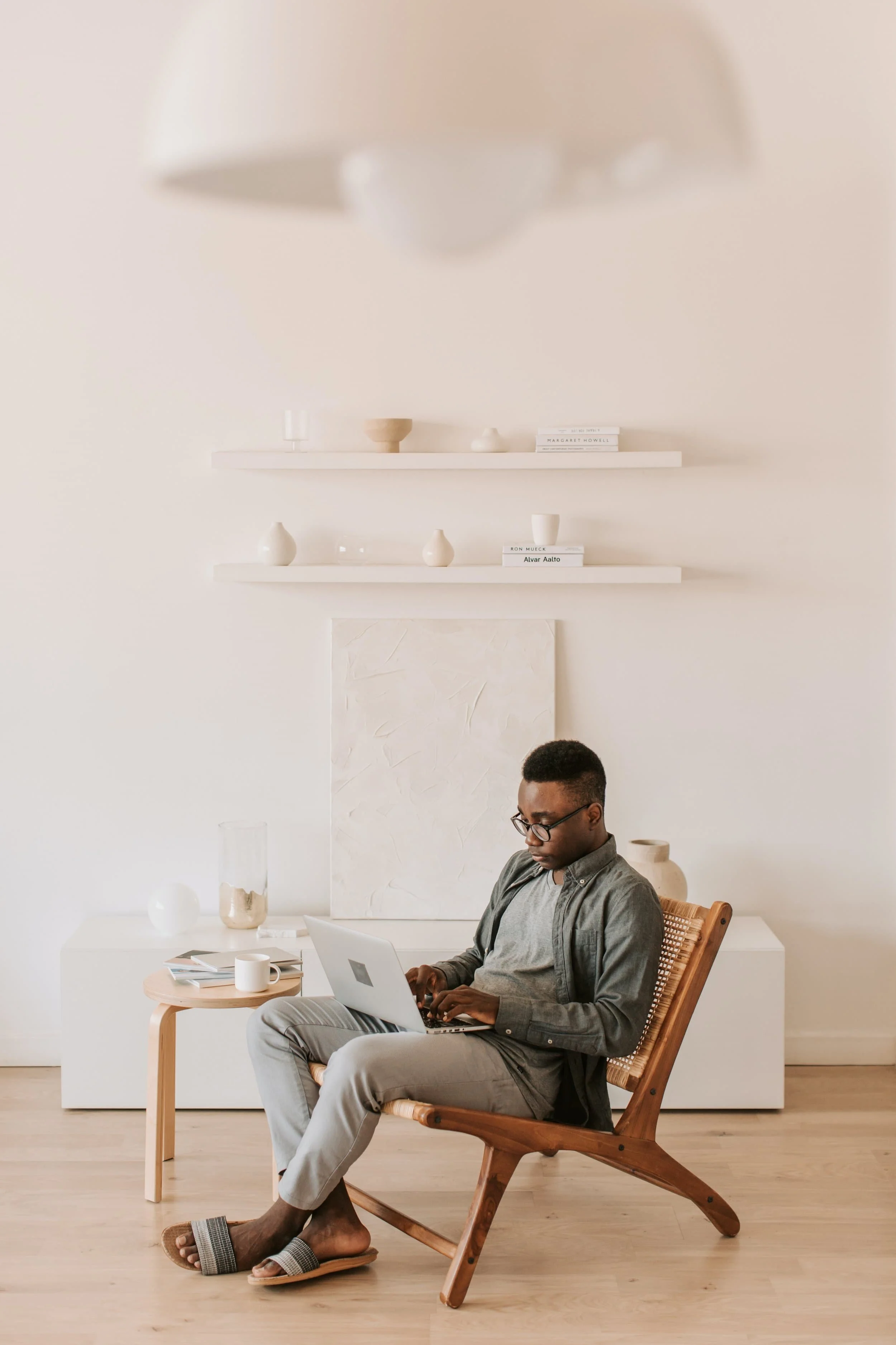 Man sitting in a wooden chair using a laptop in a minimalist, neutral-colored room with white walls and wooden floors.