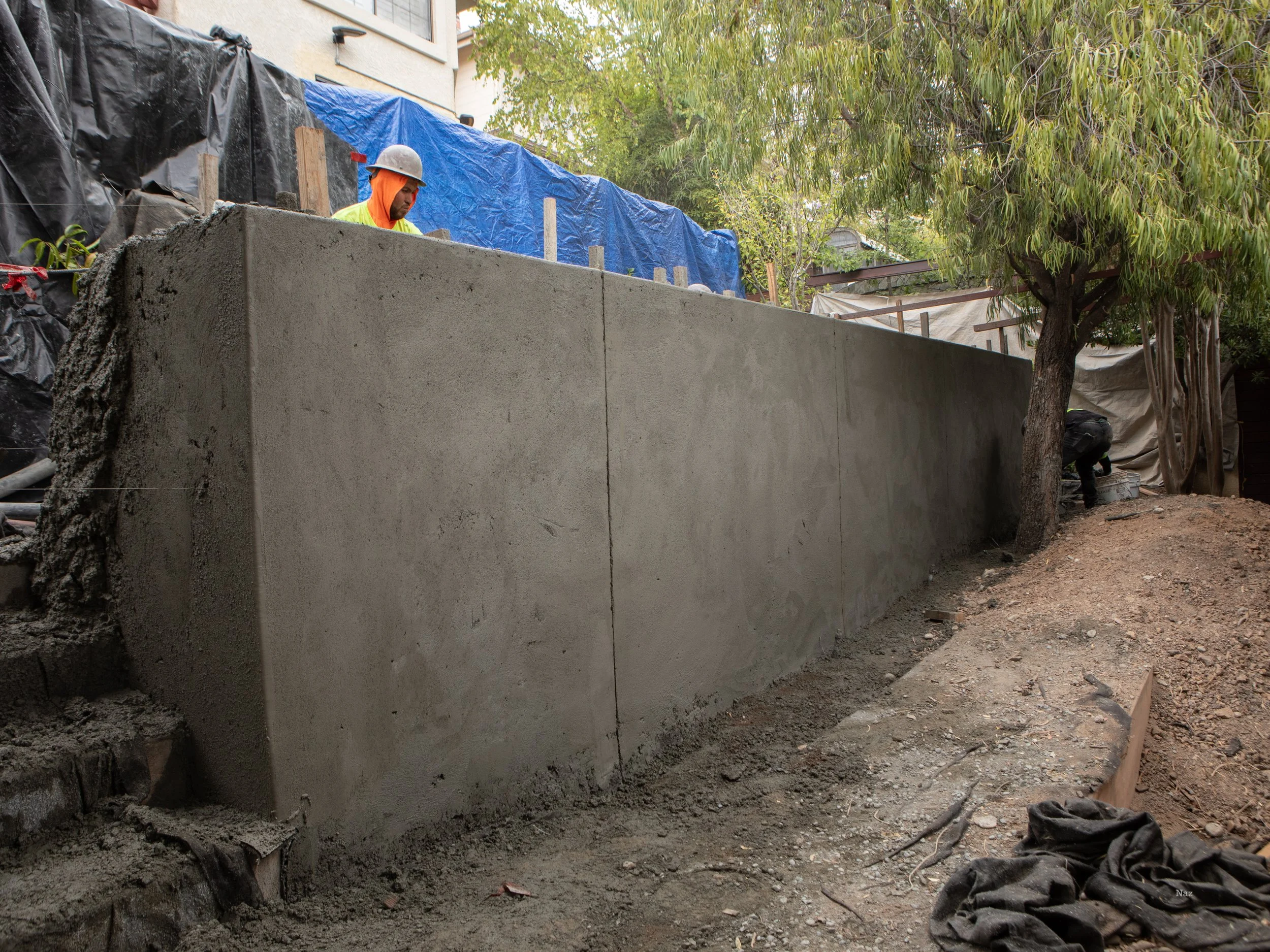 Construction workers building a concrete wall outdoors, surrounded by dirt, trees, and construction materials.