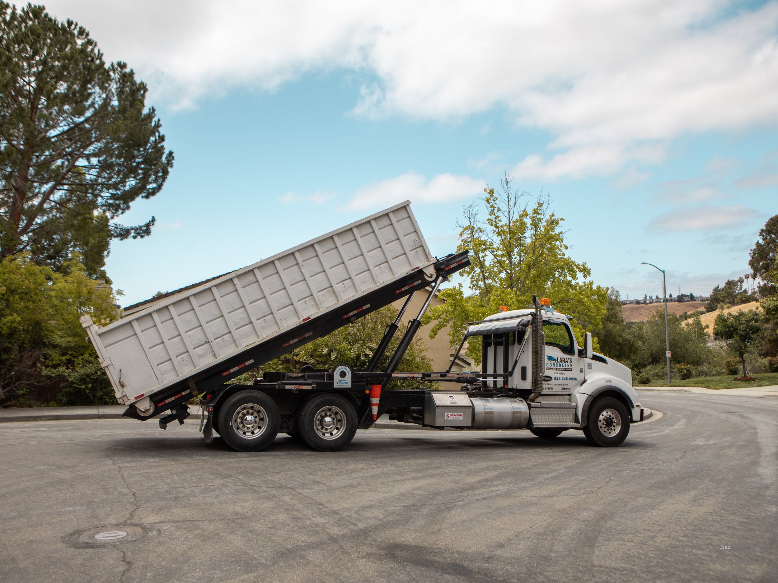 White dump truck with raised bed parked on a street with trees and hills in the background.