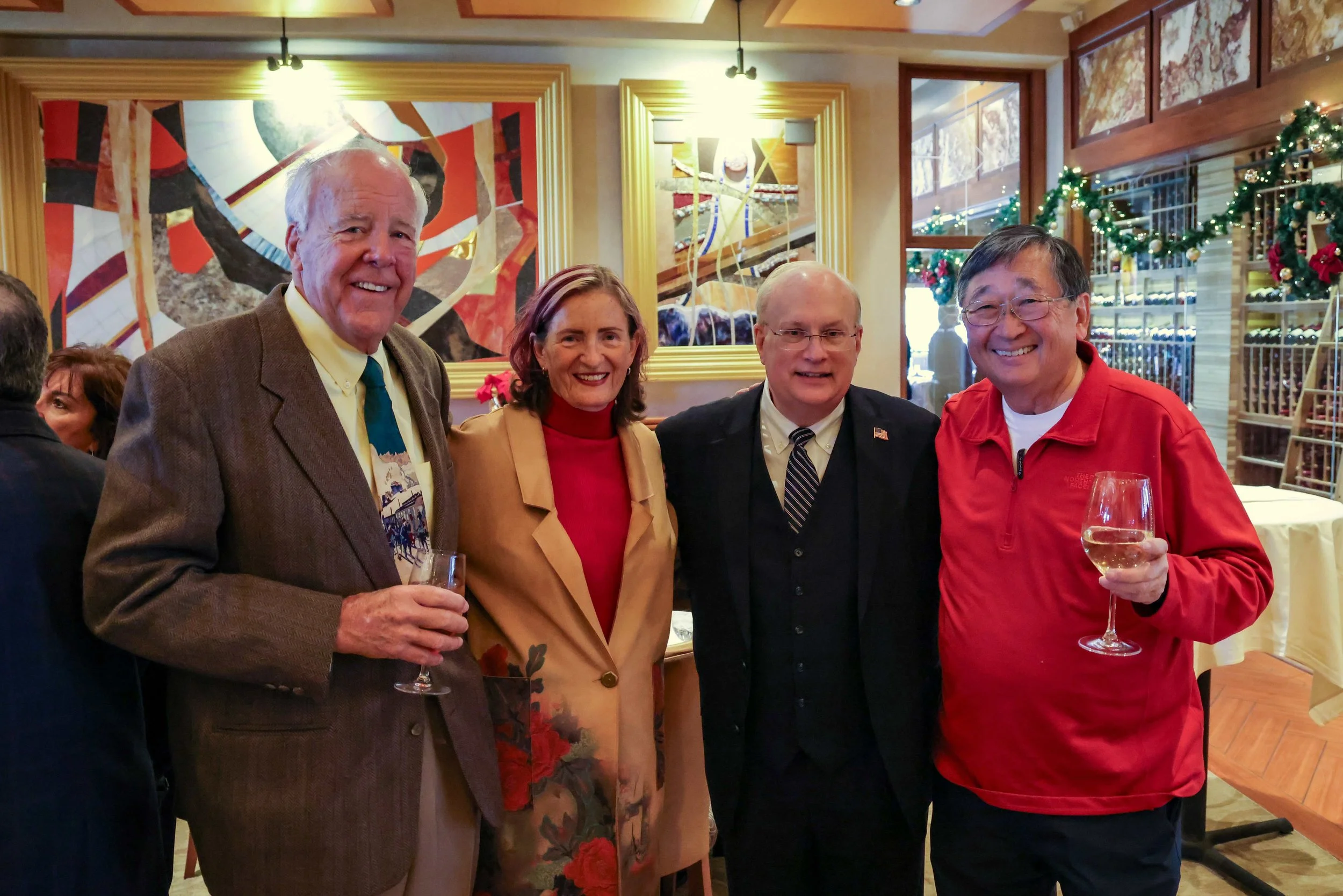 Group of four people smiling and holding glasses of white wine at a holiday gathering in a restaurant decorated with Christmas greenery and lights.
