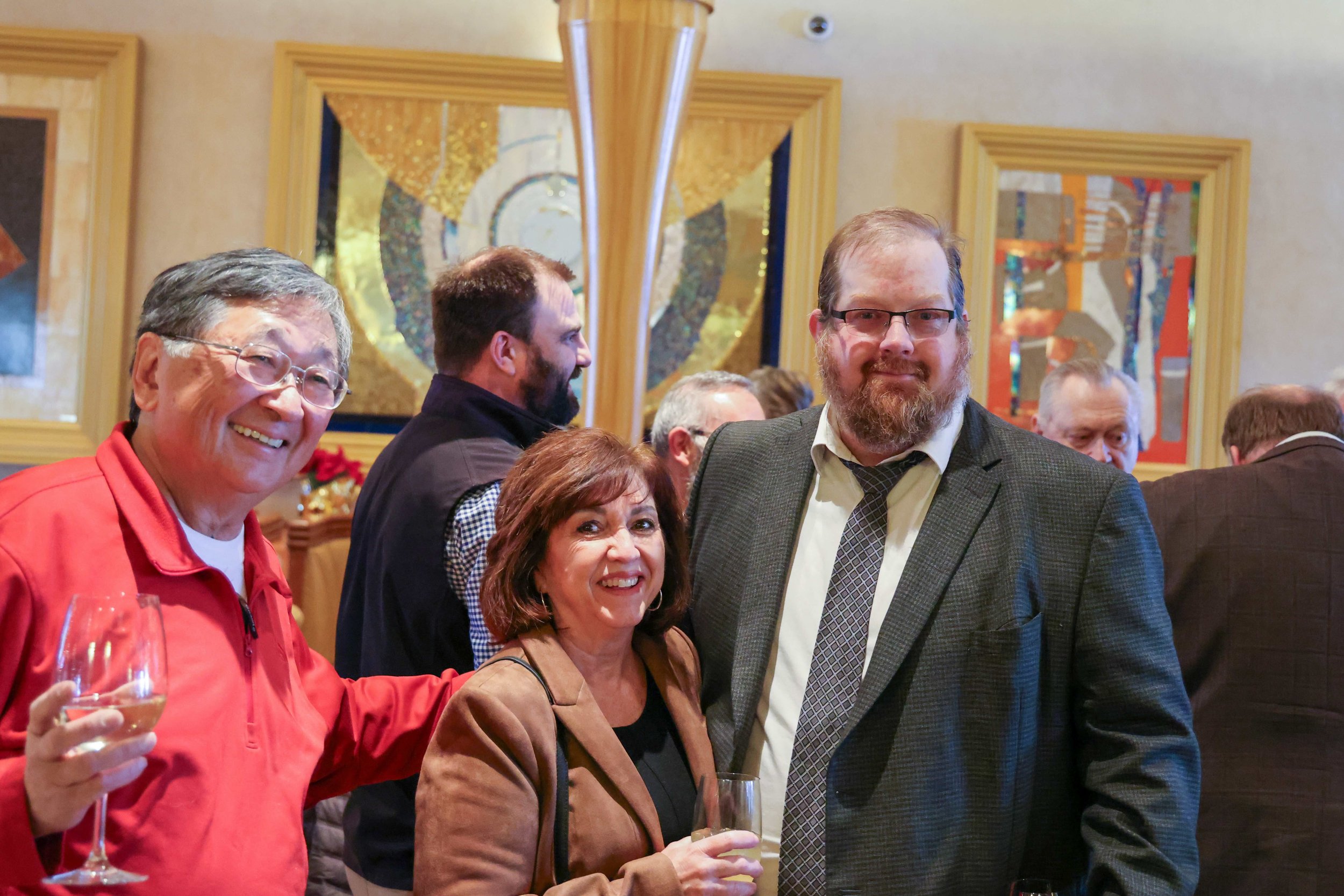 A group of people at a social event, with three individuals in the foreground smiling and holding drinks. The background features colorful artwork and other guests mingling.