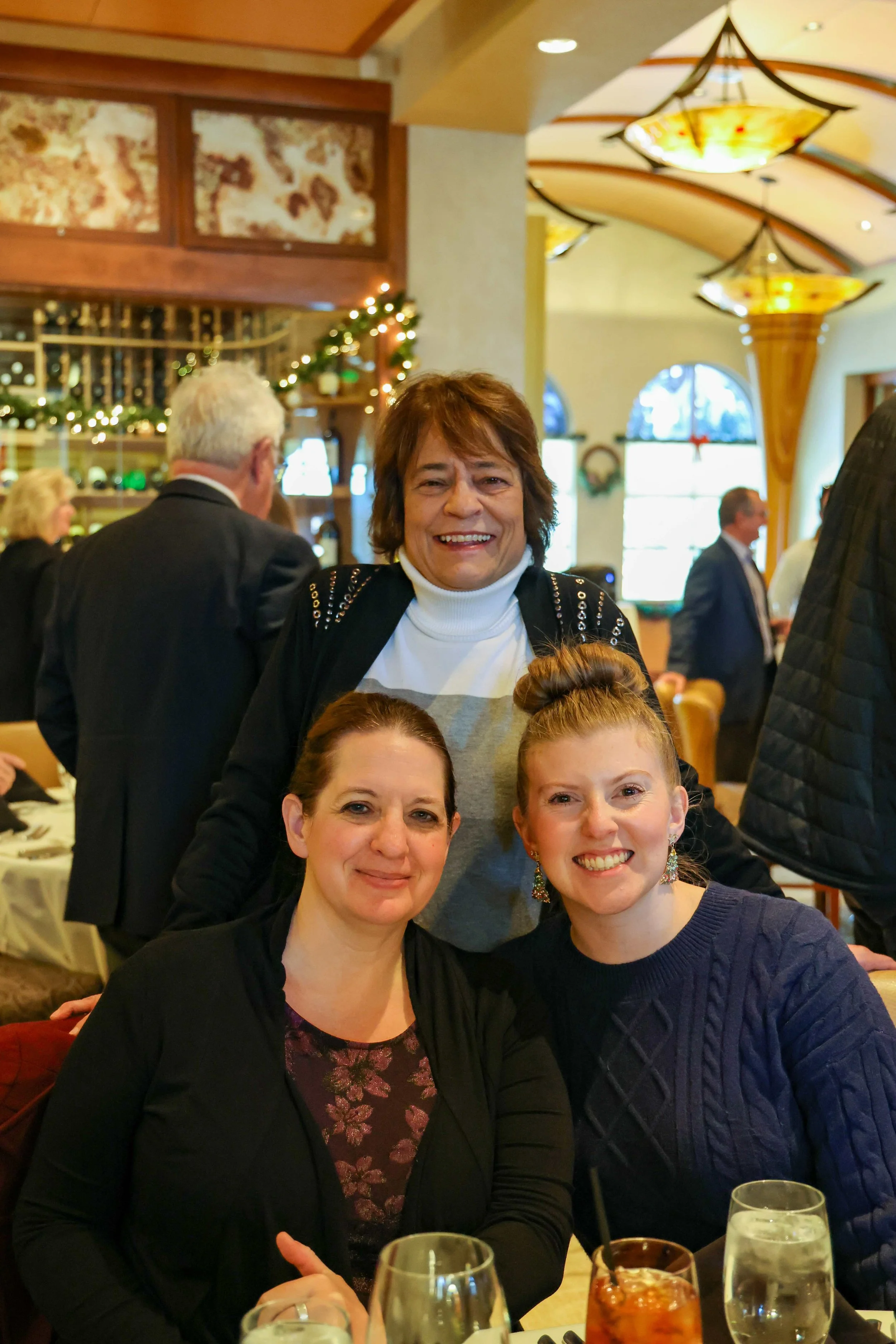 Three women smiling at a restaurant, with two seated at a table and one standing behind them, in a warmly decorated dining area.