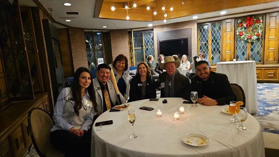 A group of six people sitting around a round table at a formal event, with some drinks and a plate of food, in a decorated room with Christmas wreath and warm lighting.