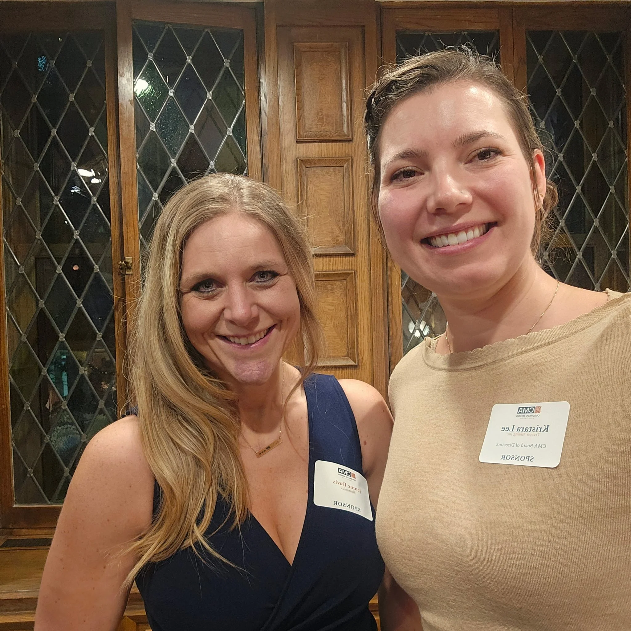 Two women smiling, taking a selfie in front of a wooden cabinet with glass doors. Both women are wearing name tags and casual clothing.