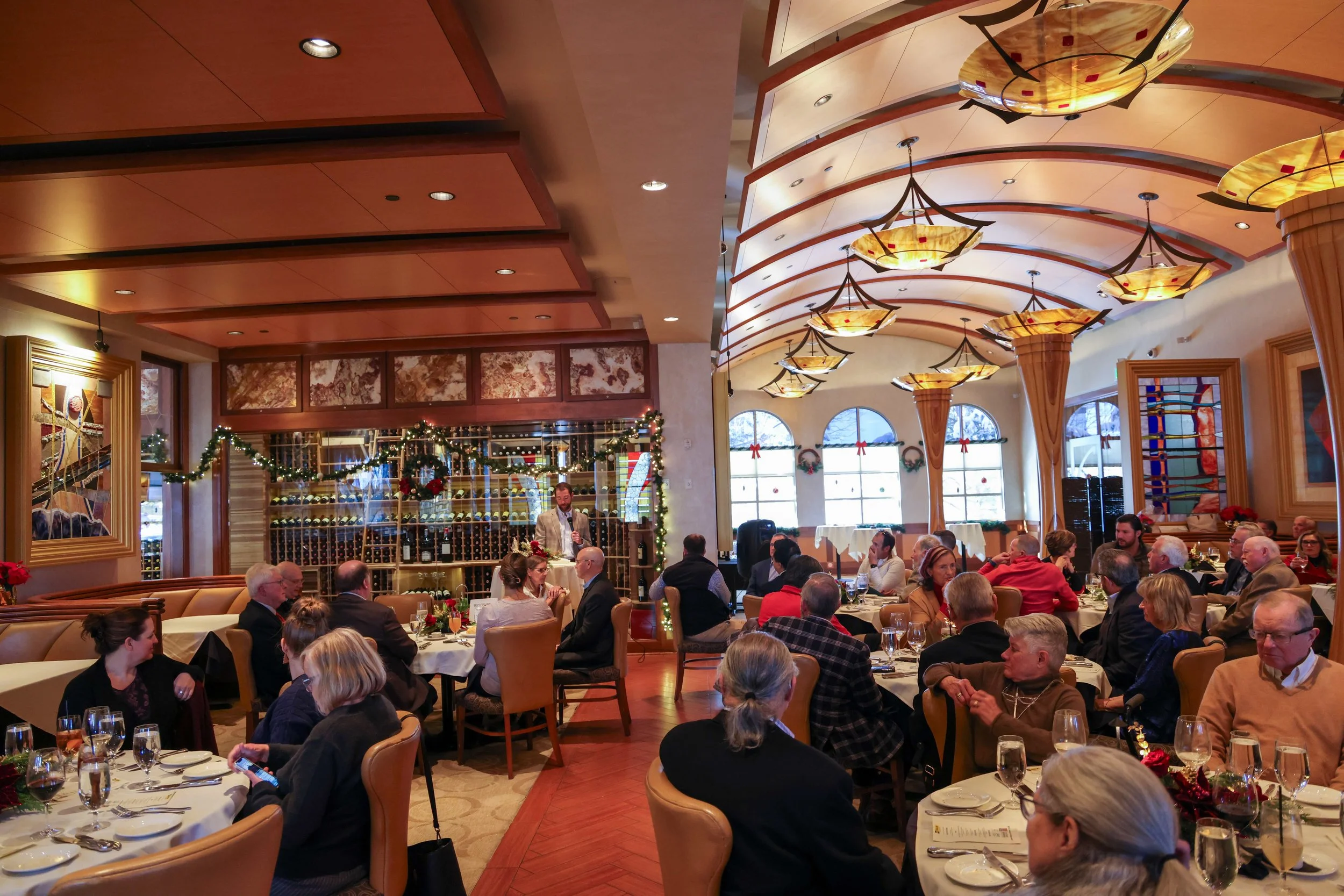 A busy restaurant decorated for Christmas, with guests seated at dining tables, some with glasses and plates, and a server speaking to one of the tables. The room has large windows with holiday wreaths, a curved ceiling with hanging lamps, and festiv