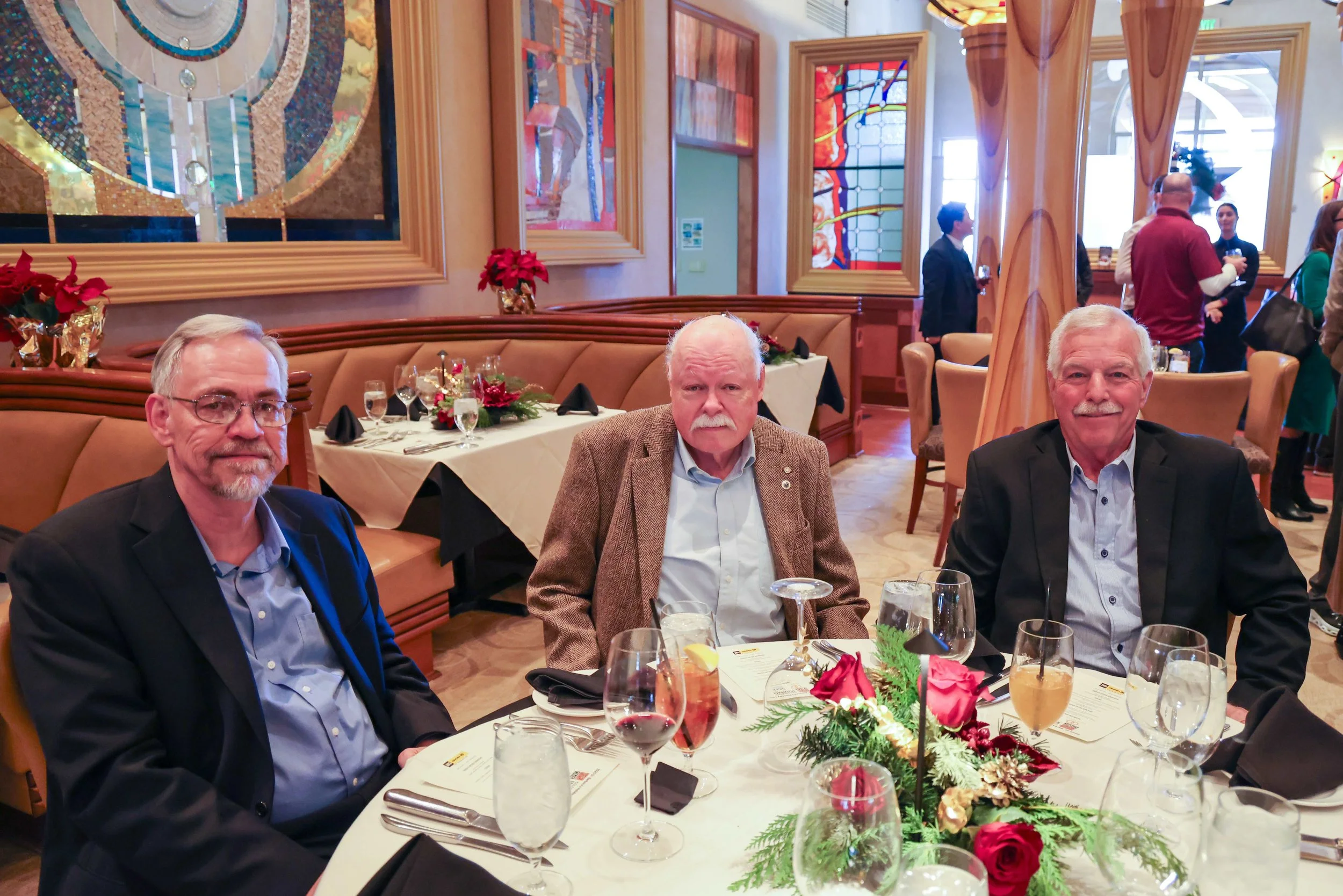 Three older men in suits sitting at a decorated Christmas table in a restaurant or banquet hall with stained glass windows and other guests in the background.